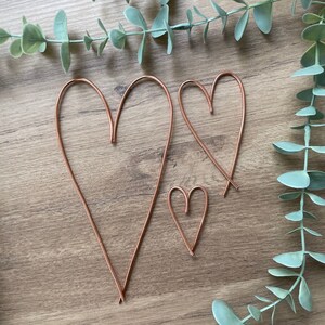 May include: Three copper wire heart shapes of varying sizes are arranged on a wooden surface, with eucalyptus leaves in the background. The hearts are simple, minimalist designs, perfect for home decor or crafting projects.