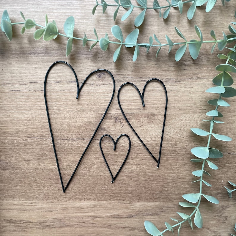 May include: Three black wire heart decorations of varying sizes are arranged on a wooden surface, with eucalyptus greenery in the background. The hearts are simple in design, perfect for wall decor or crafting projects.
