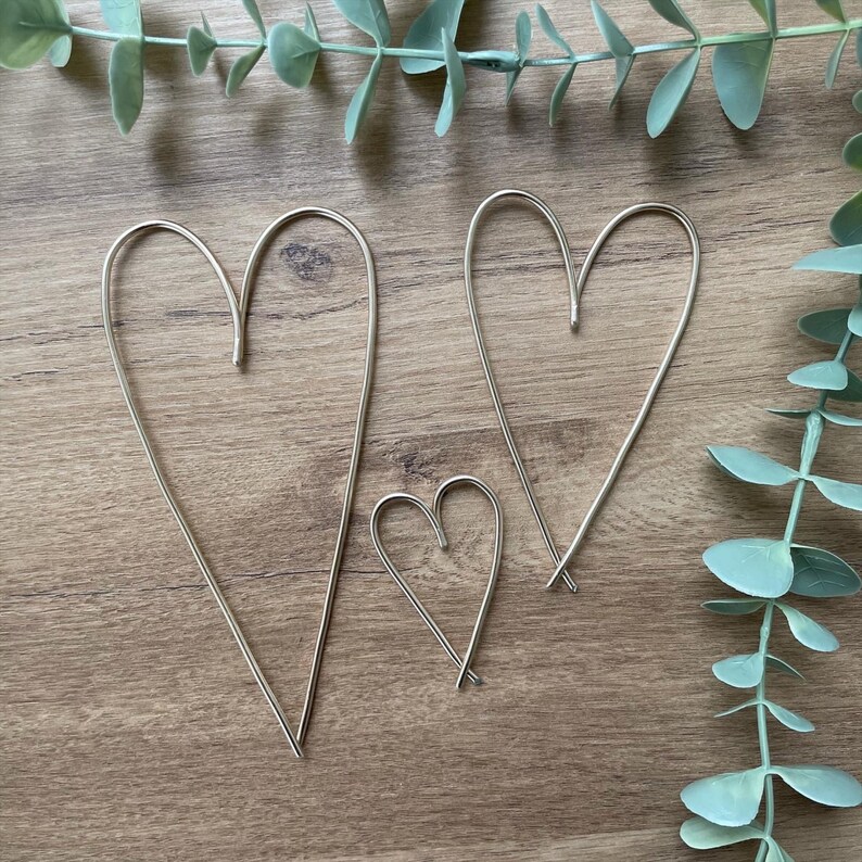 May include: Three silver-colored wire heart decorations of varying sizes. The hearts are arranged on a wooden surface, with eucalyptus leaves in the background. The largest heart is approximately 10 inches tall.