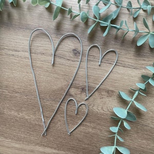 May include: Three silver wire heart shapes of varying sizes are arranged on a wooden surface, with eucalyptus greenery in the background. The hearts are simple, elegant, and likely handmade, perfect for decor or crafts.