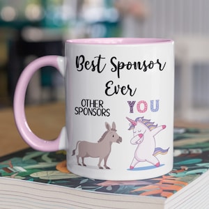 May include: White ceramic mug with a pink rim and handle. The mug features a black and white illustration of a unicorn doing the dab and a donkey. The text on the mug reads "Best Sponsor Ever" and "Other Sponsors You".