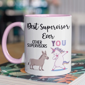 May include: White ceramic mug with a pink rim and handle. The mug features a black and white graphic of a unicorn doing the dab and a donkey. The text on the mug reads "Best Supervisor Ever Other Supervisors You".
