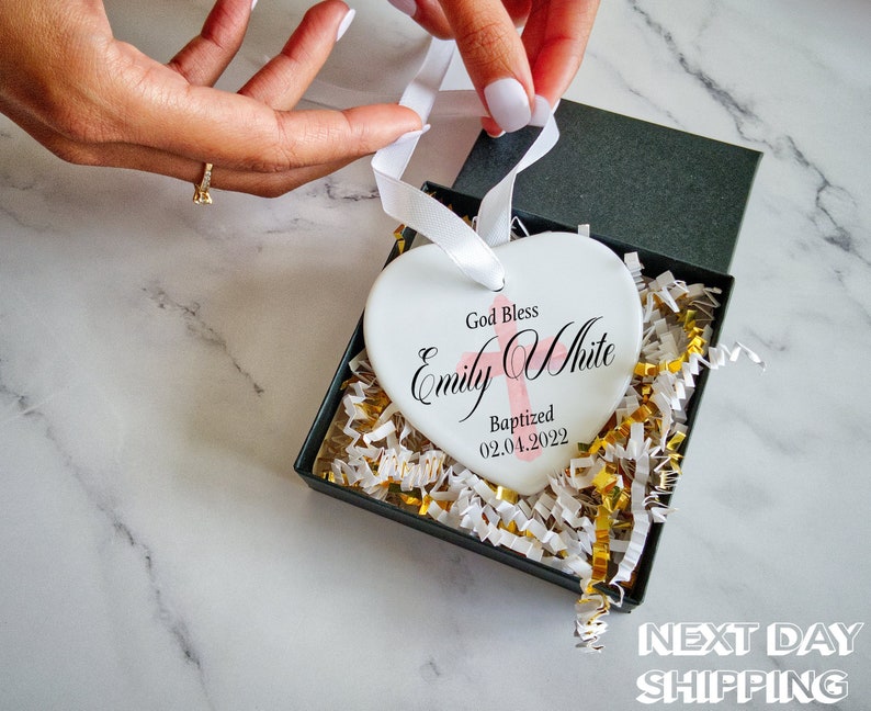 Pu&ograve; includere: Un ornamento bianco a forma di cuore con la scritta "God Bless Emily White Baptized 02.04.2022" in corsivo nero e una croce rosa. L'ornamento &egrave; in una confezione regalo nera con imbottitura bianca e oro, legato con un nastro bianco.