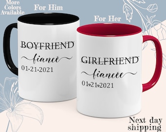 May include: Two white ceramic mugs with black and red accents. The mug on the left has the text "BOYFRIEND fiancé 01-21-2021" and the mug on the right has the text "GIRLFRIEND fiancée 01-21-2021".