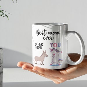 May include: White ceramic coffee mug with a black and white illustration of a unicorn and a donkey. The text on the mug reads "Best mom ever" with "Other moms" next to the donkey and "You" next to the unicorn.