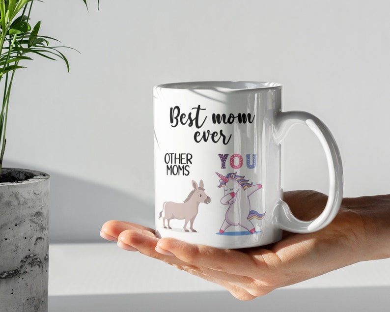 May include: White ceramic coffee mug with a black and white illustration of a unicorn and a donkey. The text on the mug reads "Best mom ever" with "Other moms" next to the donkey and "You" next to the unicorn.