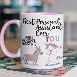 May include: White ceramic mug with a pink rim and handle. The mug has a black inscription that reads "Best Personal Assistant Ever You".  A cartoon unicorn and donkey are depicted on the mug.