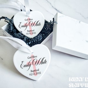 Pu&ograve; includere: Due ornamenti bianchi a forma di cuore con la scritta "God Bless Emily White Baptized 02.04.2022" in nero e rosa. Gli ornamenti sono in una scatola regalo bianca con imbottitura nera e nastro bianco. Sfondo marmoreo.