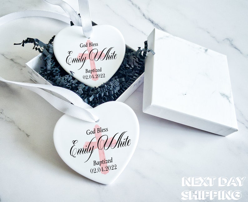 Pu&ograve; includere: Due ornamenti bianchi a forma di cuore con la scritta "God Bless Emily White Baptized 02.04.2022" in nero e rosa. Gli ornamenti sono in una scatola regalo bianca con imbottitura nera e nastro bianco. Sfondo marmoreo.
