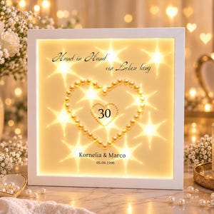 Könnte beinhalten: Ein quadratischer weißer Schattenfugenrahmen mit warmer LED-Innenbeleuchtung. In der Mitte befindet sich ein Herz aus cremefarbenen Perlen, das die Zahl 30 umschließt. Der Text lautet: Hand in Hand ein Leben lang, Kornelia & Marco, 05.09.1996.