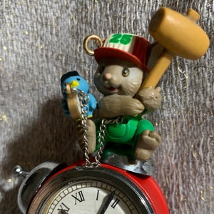 May include: A red and silver alarm clock with a white face and black Roman numerals. A small figurine of a cartoon rabbit wearing a hat and holding a mallet sits atop the clock. A blue bird figurine hangs from a chain.
