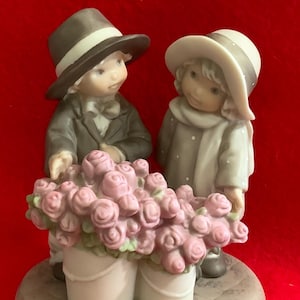 Puede incluir: Figura de porcelana que representa a dos niños, un niño y una niña, junto a un gran ramo de rosas rosas. El niño lleva un traje y sombrero, la niña un abrigo y sombrero. La figura está sobre un fondo rojo.