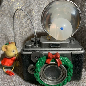 May include: A silver and black camera ornament with a green wreath and red bow around the lens. A small mouse in a red dress is holding a wire. The camera has a flash with a white bulb.