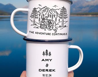 May include: Two white enamel camping mugs stacked on top of each other. The top mug has a black illustration of a mountain scene with the text "THE ADVENTURE CONTINUES". The bottom mug has the names "AMY & DEREK" and the date "10.12.24".