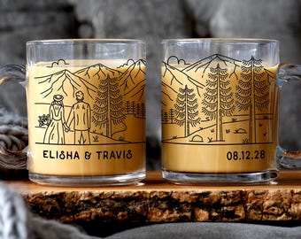 May include: Two clear glass mugs with handles, each filled with a light brown liquid. The mugs feature black line art of a mountain scene with trees. One mug has the text "ELISHA & TRAVIS", the other "08.12.28".