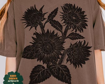 May include: A brown short-sleeve t-shirt featuring a large black graphic print of three sunflowers with detailed petals and leaves. The design is centered on the chest area. A small green clover-shaped emblem with the text HAND DRAWN ARTWORK is visible.