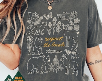 May include: A charcoal gray short-sleeve t-shirt featuring a white line-art graphic of various forest animals, including a bear, deer, squirrel, and goat, alongside plants and the text "respect the locals" printed in a gold script font.