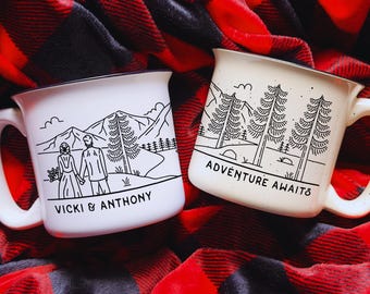 May include: Two white ceramic mugs with black outlines. One mug features a couple standing in front of a mountain range with the text "Vicki & Anthony". The other mug features a mountain range with trees and the text "Adventure Awaits".