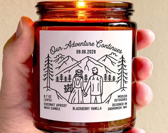 May include: A lit, brown glass candle jar with a white label featuring a mountain scene and the text "Our Adventure Continues 09.06.2028." The candle is 8.7 oz (247g) and scented with coconut apricot wax and blackberry vanilla.
