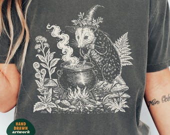 May include: A charcoal gray t-shirt featuring a white graphic illustration of an opossum wearing a witch hat, stirring a steaming cauldron over a fire. The design includes ferns and mushrooms. A small green badge reads HAND DRAWN ARTWORK.
