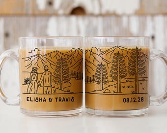 May include: Two clear glass mugs with a mountain and forest design. One mug features a couple holding hands with the text "ELISHA & TRAVIS". The other mug has the date "08.12.28". Both mugs are filled with a light brown liquid.