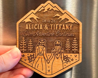 May include: A wooden engraved sign with a mountain scene and two figures walking. The sign reads "08.16.26 Alicia & Tiffany Our Adventure Continues".