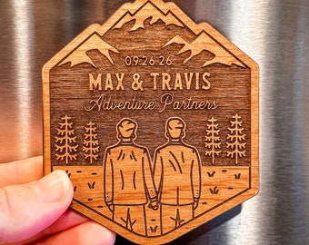 May include: A wooden engraved sign with a mountain scene, two figures holding hands, and the text "09.26.26 MAX & TRAVIS Adventure Partners".