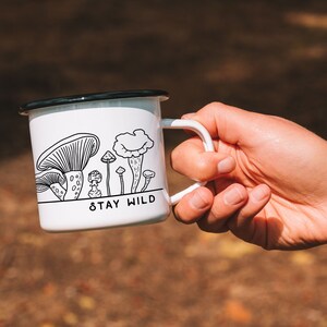 May include: White enamel mug with a black rim and handle. The mug has a black and white illustration of mushrooms and the text "Stay Wild".