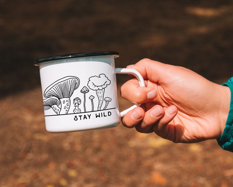 May include: White enamel mug with a black rim and handle. The mug has a black and white illustration of mushrooms and the text "Stay Wild".