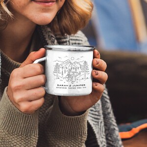 May include: White enamel camping mug with a black rim and handle. The mug features a black and white line drawing of a mountain scene with trees, a river, and two figures. The text "SARAH & JUNIPER ADVENTURING TOGETHER SINCE 2016" is printed on the mug.