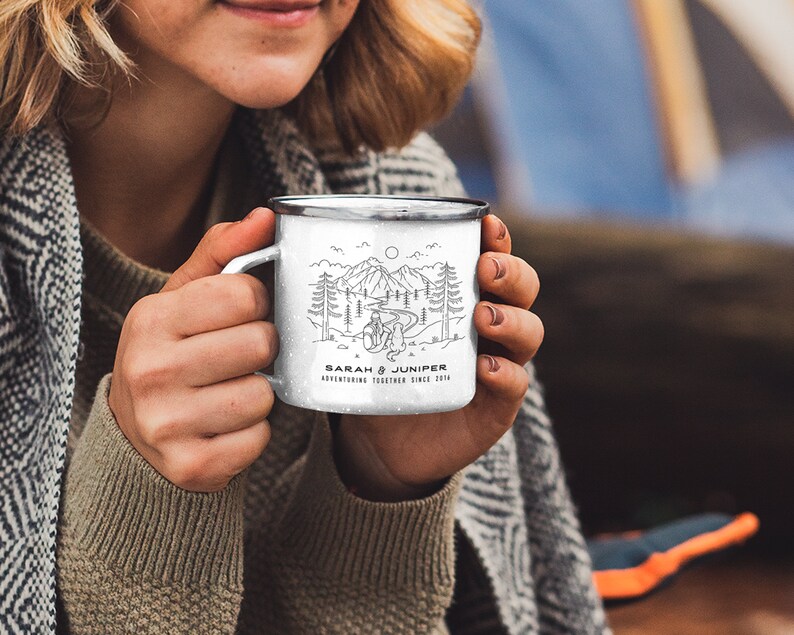 May include: White enamel camping mug with a black rim and handle. The mug features a black and white line drawing of a mountain scene with trees, a river, and two figures. The text "SARAH & JUNIPER ADVENTURING TOGETHER SINCE 2016" is printed on the mug.