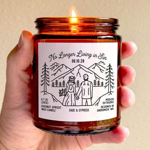 Könnte beinhalten: Eine brennende, bernsteinfarbene Glas-Kerze, die in einer Hand gehalten wird. Die Kerze hat ein weißes Etikett mit schwarzem Text und Illustrationen eines Paares, Bergen und Bäumen. Das Etikett lautet "No Longer Living in Sin" und "06.10.28". Die Kerze ist 247g.