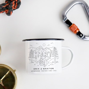 May include: White enamel camping mug with a black outline. The mug features a black and white illustration of a person and a dog hiking in a mountain landscape. The text "Erik & Baxter Adventuring Together Since 2016" is printed on the bottom of the mug.