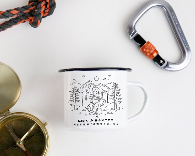 May include: White enamel camping mug with a black outline. The mug features a black and white illustration of a person and a dog hiking in a mountain landscape. The text "Erik & Baxter Adventuring Together Since 2016" is printed on the bottom of the mug.