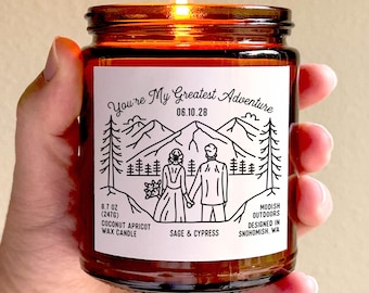 May include: A lit, amber-colored glass jar candle held in a hand. The label reads "You're My Greatest Adventure" with a mountain and couple illustration. It is a 8.7 oz (247g) coconut apricot wax candle with sage and cypress scent.