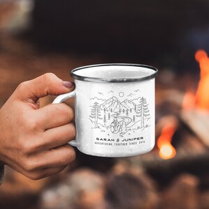 May include: White enamel camping mug with a black rim and handle. The mug features a black and white illustration of a mountain scene with the text "SARAH & JUNIPER ADVENTURING TOGETHER SINCE 2014". The mug is held by a person.