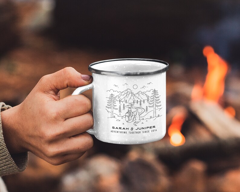 May include: White enamel camping mug with a black rim and handle. The mug features a black and white illustration of a mountain scene with the text "SARAH & JUNIPER ADVENTURING TOGETHER SINCE 2014". The mug is held by a person.