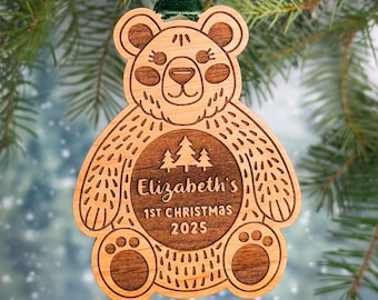 May include: A wooden bear ornament with a brown stain. The bear is holding a circle with the text "Elizabeth's 1st Christmas 2025" and three small pine trees.