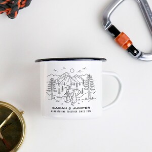 May include: White enamel camping mug with a black rim and handle. The mug features a black and white illustration of a mountain scene with the text "SARAH & JUNIPER ADVENTURING TOGETHER SINCE 2014". A compass and carabiner are in the background.