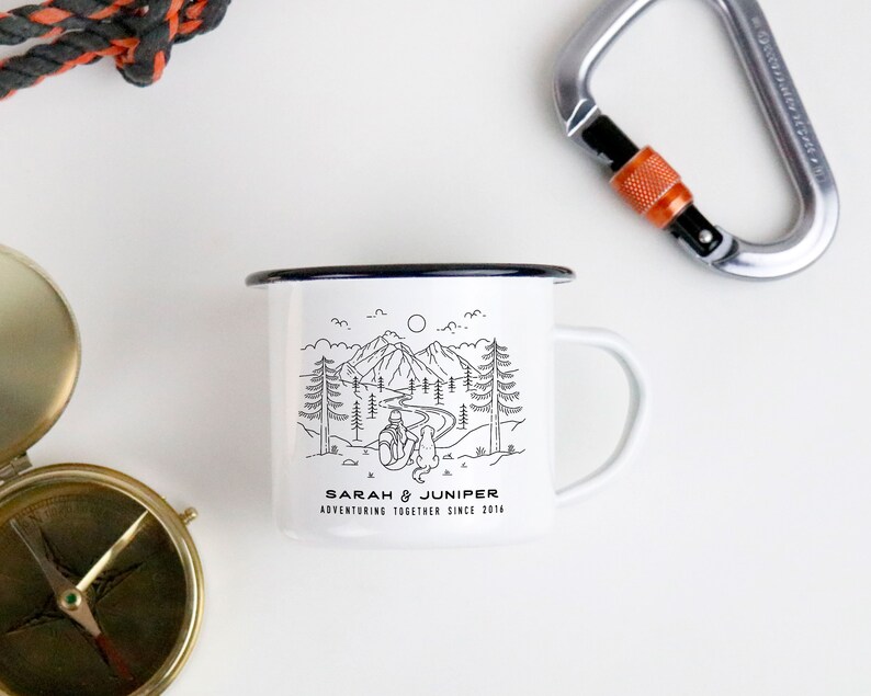May include: White enamel camping mug with a black rim and handle. The mug features a black and white illustration of a mountain scene with the text "SARAH & JUNIPER ADVENTURING TOGETHER SINCE 2014". A compass and carabiner are in the background.