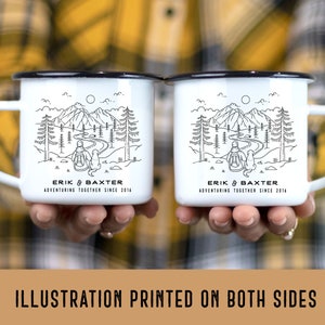 May include: Two white enamel mugs with black rims. Each mug features a black line drawing of a person and a dog walking through a forest with mountains in the background. The text "ERIK & BAXTER ADVENTURING TOGETHER SINCE 2016" is printed on the bottom of each mug.