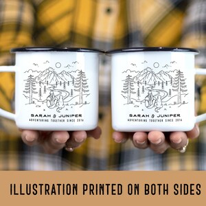 May include: Two white enamel mugs with black rims and handles, each featuring a black and white mountain and forest illustration. The mugs have the text "SARAH & JUNIPER ADVENTURING TOGETHER SINCE 2014". The mugs are held in front of a plaid shirt.