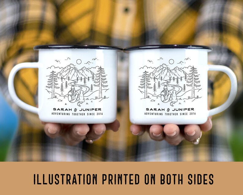May include: Two white enamel mugs with black rims and handles, each featuring a black and white mountain and forest illustration. The mugs have the text "SARAH & JUNIPER ADVENTURING TOGETHER SINCE 2014". The mugs are held in front of a plaid shirt.