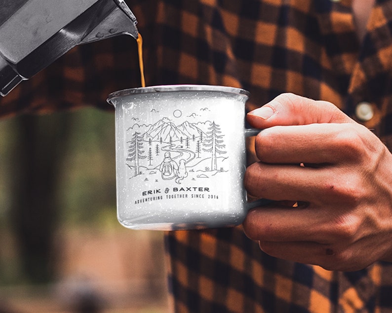 May include: A white enamel camping mug with a black line drawing of a person and a dog hiking in the mountains. The mug has the text "Erik & Baxter Adventuring Together Since 2014" printed on the bottom.