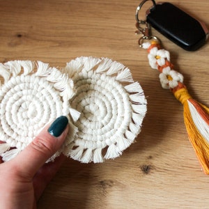 May include: Two off-white macrame coasters with a spiral design and fringe detail. A matching keychain with white daisy accents and a tassel in orange, yellow, and white is also visible. The items are on a wooden surface.