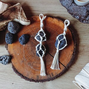 Puede incluir: Dos adornos colgantes de macramé con piedras de lava negra. Los adornos están hechos con cordón blanco y tienen un borla en la parte inferior. Los adornos se muestran en una rodaja de madera.
