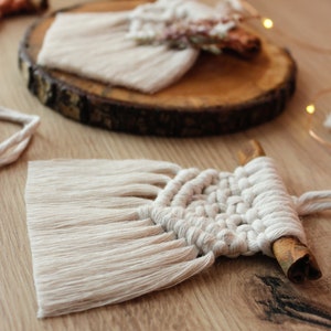 May include: A white macrame wall hanging with a cinnamon stick attached to the bottom. The wall hanging is made with a knotting technique and has a fringe on the bottom.
