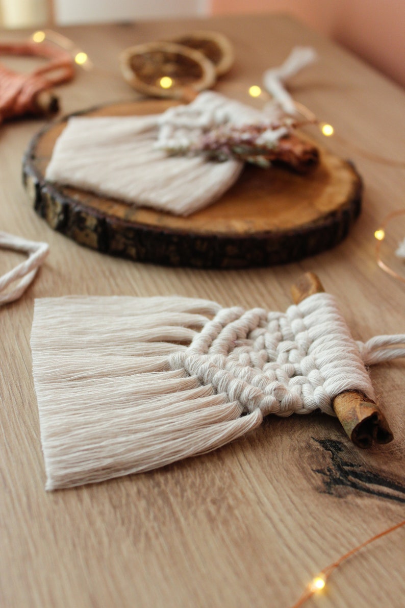 May include: A white macrame wall hanging with a cinnamon stick attached to the bottom. The wall hanging is made with a knotting technique and has a fringe on the bottom.