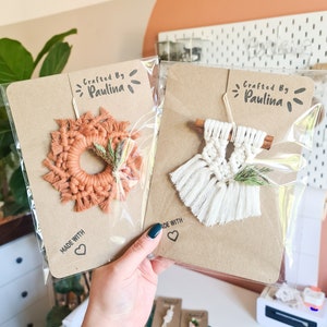 May include: Two macrame wall hangings with a rustic brown background. The top left wall hanging is a round orange macrame wreath with a small sprig of dried greenery. The top right wall hanging is a white macrame wall hanging with a cinnamon stick and a sprig of dried greenery. Both hangings have a "Crafted by Paulina" label and a "Made with Love" label.
