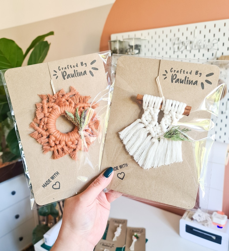 May include: Two macrame wall hangings with a rustic brown background. The top left wall hanging is a round orange macrame wreath with a small sprig of dried greenery. The top right wall hanging is a white macrame wall hanging with a cinnamon stick and a sprig of dried greenery. Both hangings have a "Crafted by Paulina" label and a "Made with Love" label.